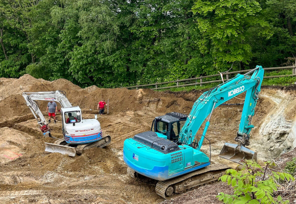 Ein blauer Kobelco-Bagger und ein weißer Takeuchi-Minibagger arbeiten auf einer Baustelle mit Erdaushub, umgeben von Bäumen.