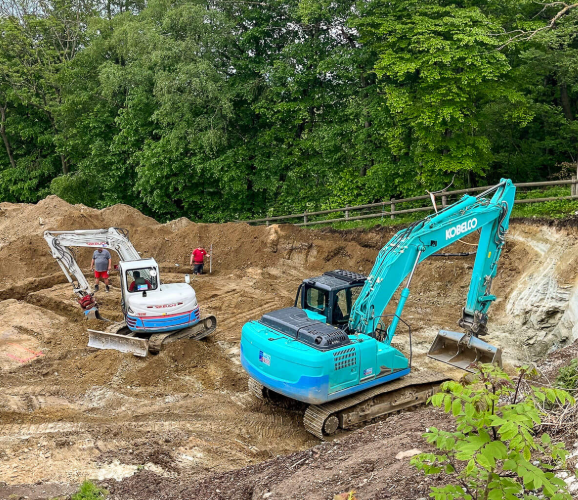 Ein blauer Kobelco-Bagger und ein weißer Takeuchi-Minibagger arbeiten auf einer Baustelle mit Erdaushub, umgeben von Bäumen.
