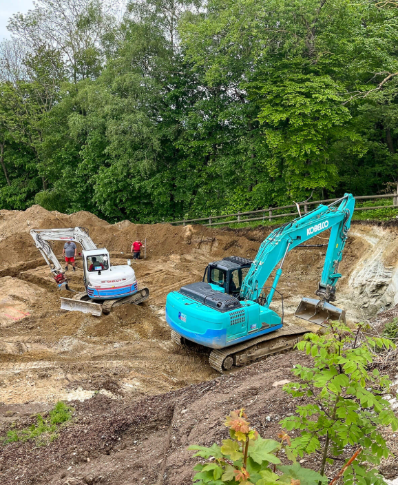 Ein blauer Kobelco-Bagger und ein weißer Takeuchi-Minibagger arbeiten auf einer Baustelle mit Erdaushub, umgeben von Bäumen.