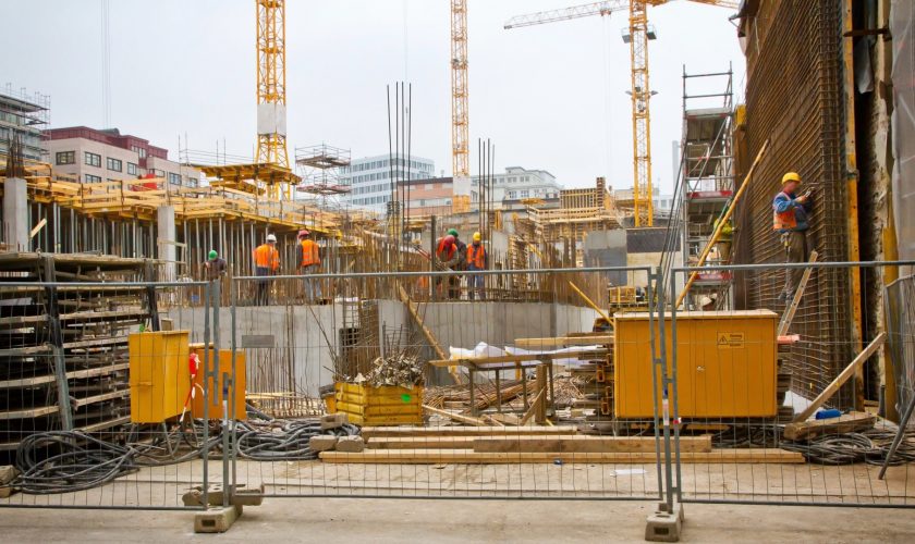Baustelle mit Bauzaun, Kränen, Baumaterial und mehreren Arbeitern im Rohbau