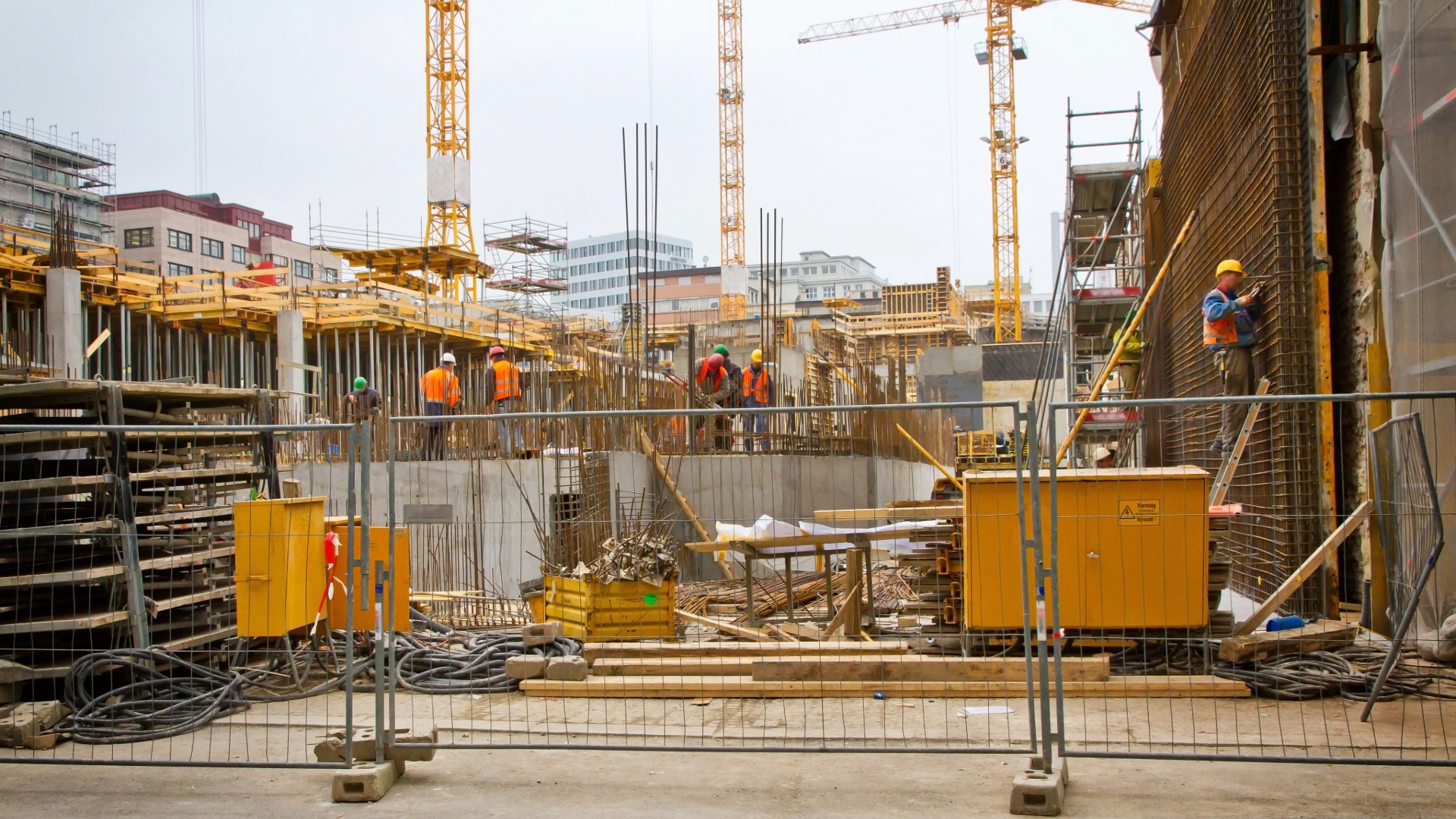 Baustelle mit Bauzaun, Kränen, Baumaterial und mehreren Arbeitern im Rohbau